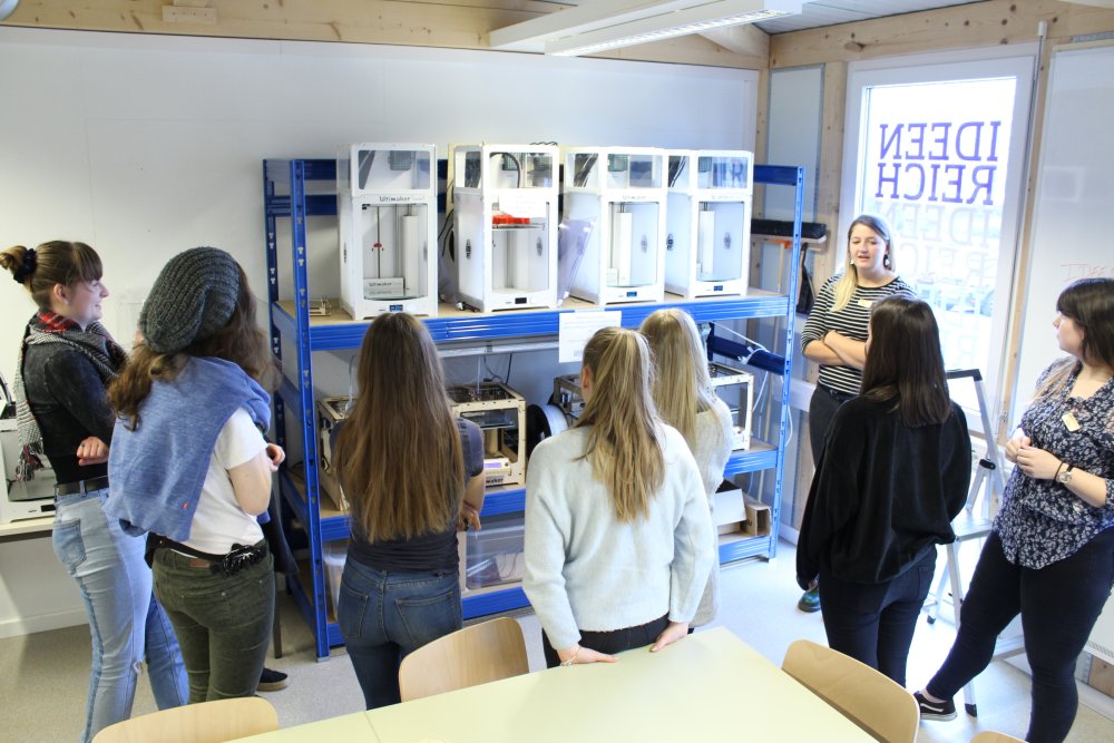 Eine Gruppe von Mädchen steht vor einem Schwerlastregal mit verschiedenen 3-D-Druckern, vor dem eine Frau steht und mit den Mädchen spricht.