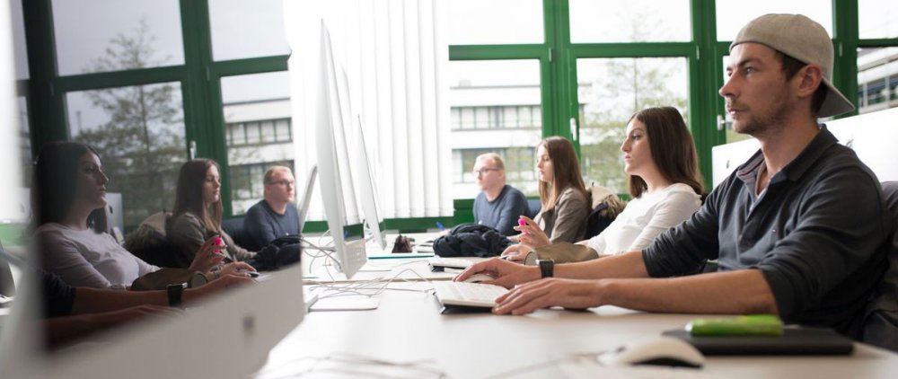 Studierende arbeiten im Seminarraum an Computern. Sie werden dabei in einem Bildschirm gespiegelt.