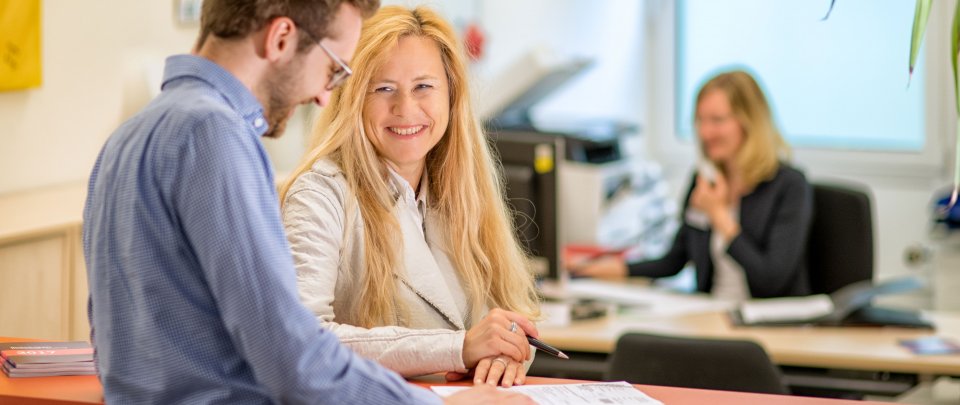 Mitarbeiterin des Studierendenservice im Gespräch mit einem Studenten. Im Hintergrund arbeitet eine weitere Mitarbeiterin am Schreibtisch.