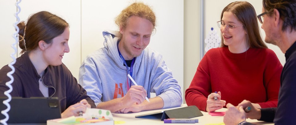 Studierende und ein Dozent stehen um einen Tsich. Sie halten Stifte, einer macht Notizen auf dem Tablet.