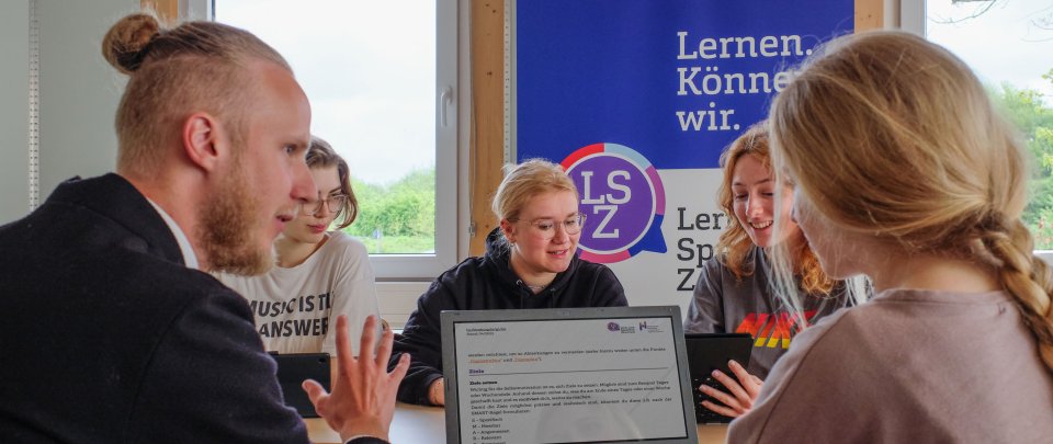 Gruppe Studierender um einen Tisch versammelt, sie schauen auf einen Laptop oder sidn im Gespräch. Hinter ihnen ein Roll Up mit LSZ-Aufdruck.