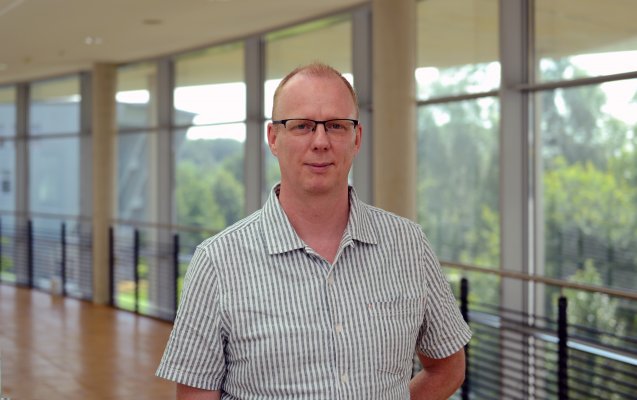 Porträtfoto: Hendrik van der Sluis im audimax. Er trägt ein weßes Hemd mit grauen Streifen Hinter ihm ist die Fensterfront des audimax mit Ausblick auf den grünen Campus und der Parkettboden zu sehen.