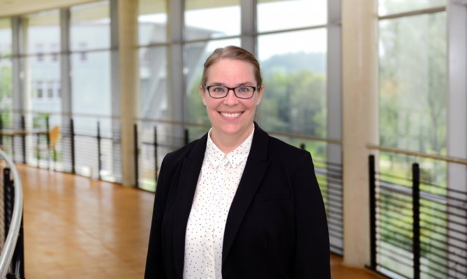 Porträtfoto: Beatrice Podtschaske im audimax. Sie trägt einen dunklen Blazer über einer hellen Bluse. Hinter ihr ist die Fensterfront des audimax mit Ausblick auf den grünen Campus und der Parkettboden zu sehen.