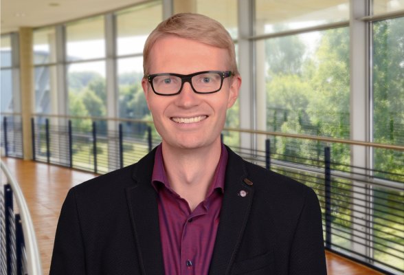 Porträtfoto: Claus Hartmann im audimax. Er trägt kurze Haare und eine Brille. Er lacht in die Kamera. Er trägt ein dunkles Sakko über einem weinroten Hemd.