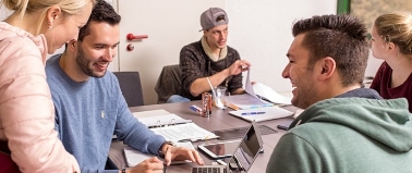 Eine Projektgruppe sitzt zusammen an einem Tisch und arbeitet zusammen an einem Projekt und hat sichtlich Spaß dabei.